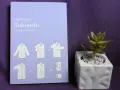 Książka "Tokimeki" Marie Kondo z instrukcją składania ubrań. Odkryj magię sprzątania i porządek w szafie. Obok mała roślinka w doniczce.