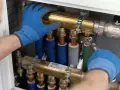 Instalator w niebieskich rękawiczkach dokręca kluczem mosiężny rozdzielacz, zapewniając prawidłowe odwodnienie domu.