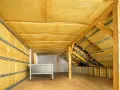 Wnętrze poddasza z widoczną wełną mineralną, która jest kluczowa, jak ocieplić dach. Drewniane belki i metalowe stelaże tworzą konstrukcję.