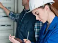 Młoda elektryczka w białym kasku i niebieskim kombinezonie notuje dane, podczas gdy kolega wskazuje na rozdzielnicę. Potwierdzenie kwalifikacji, certyfikat elektryczny w tle.