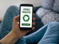 Dłoń trzyma smartfon z ekranem aplikacji zero waste. Zielony symbol recyklingu i przycisk "Learn More" zachęcają do ekologicznych działań.