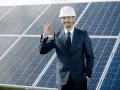 Uśmiechnięty mężczyzna w kasku i garniturze pokazuje znak OK na tle paneli fotowoltaicznych. To symbol efektywnego oddawania energii do sieci.