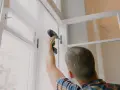 Mężczyzna przykręca zawias w ramach okna. Trwa wymiana okien, przygotowanie do montażu.