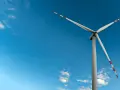 Mała elektrownia wiatrowa z białymi łopatami i czerwono-białymi końcówkami na tle błękitnego nieba z chmurami.