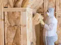 Ocieplanie wełną mineralną domu drewnianego. Pracownik w kombinezonie montuje izolację w drewnianej konstrukcji.