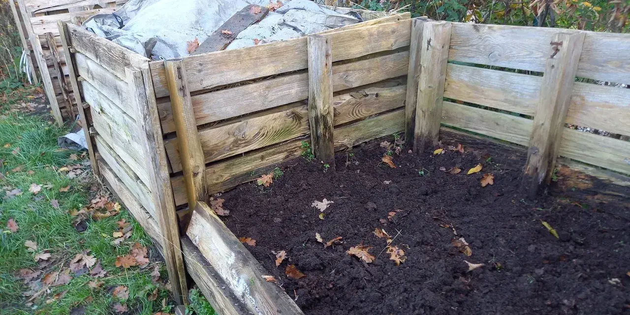 Drewniany kompostownik w ziemi, częściowo przykryty plandeką. Widać jesienne liście i trawę.