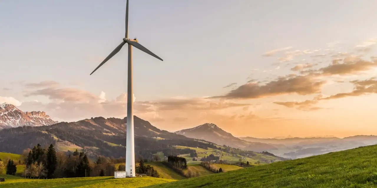 Wiatrak na zielonym wzgórzu, symbolizujący czystą energię z wiatru, na tle malowniczych gór o zachodzie słońca.