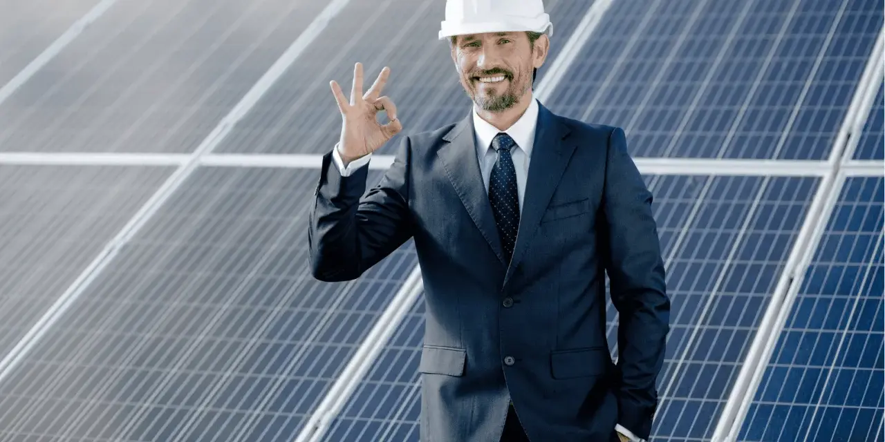Uśmiechnięty mężczyzna w kasku i garniturze pokazuje znak OK na tle paneli fotowoltaicznych. To symbol efektywnego oddawania energii do sieci.