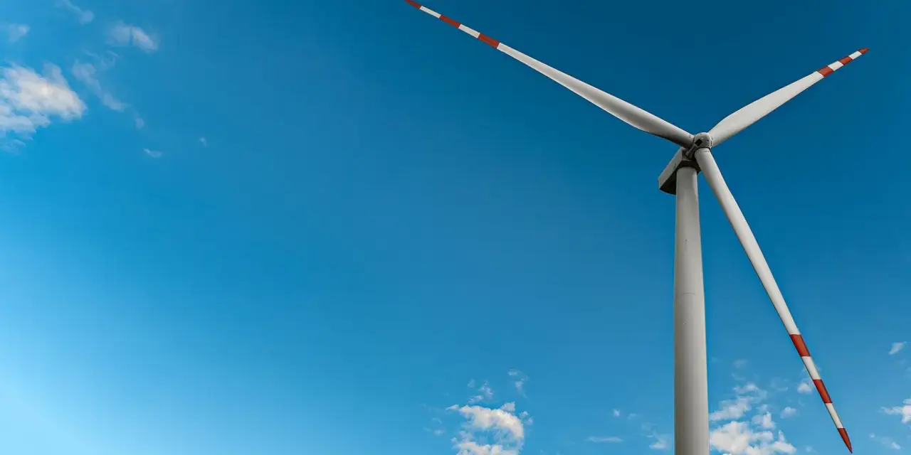 Mała elektrownia wiatrowa z białymi łopatami i czerwono-białymi końcówkami na tle błękitnego nieba z chmurami.