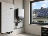 Nowoczesny magazyn energii i falownik, gotowe do instalacji najlepszej fotowoltaiki. Za oknem widać dom z panelami słonecznymi na dachu.
