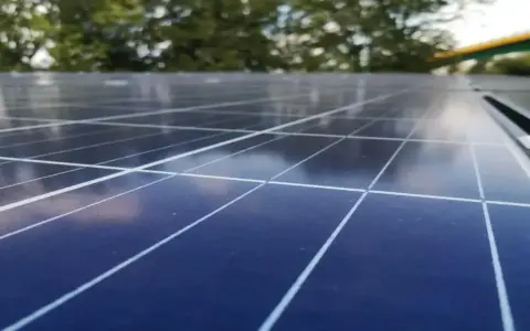 Zbliżenie na panele fotowoltaiczne, które zamieniają światło słoneczne w energię. W tle drzewa i most.