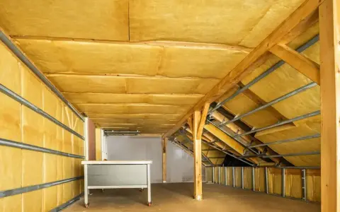 Wnętrze poddasza z widoczną wełną mineralną, która jest kluczowa, jak ocieplić dach. Drewniane belki i metalowe stelaże tworzą konstrukcję.