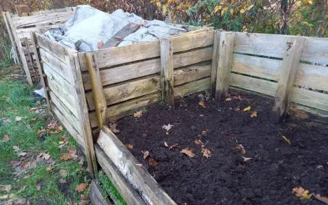 Drewniany kompostownik w ziemi, częściowo przykryty plandeką. Widać jesienne liście i trawę.