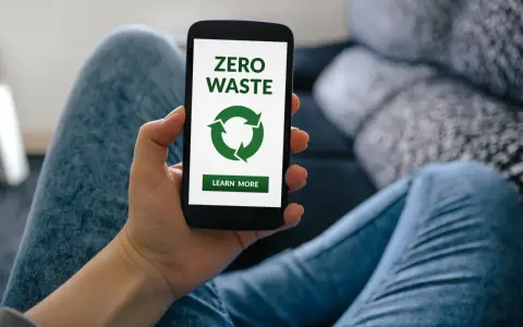 Dłoń trzyma smartfon z ekranem aplikacji zero waste. Zielony symbol recyklingu i przycisk "Learn More" zachęcają do ekologicznych działań.