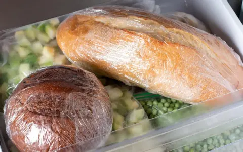 Dwa bochenki chleba, jeden jasny, drugi ciemny, owinięte folią spożywczą, czekają na zamrożenie. Obok mrożone warzywa.