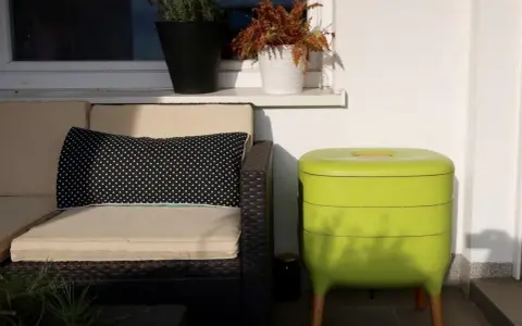 Zielony kompostownik na balkonie, obok wygodnej kanapy z poduszką w grochy.