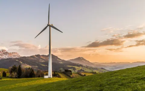 Wiatrak na zielonym wzgórzu, symbolizujący czystą energię z wiatru, na tle malowniczych gór o zachodzie słońca.