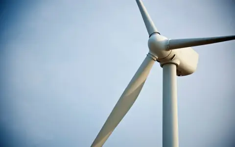 Biały wiatrak elektrownia na tle błękitnego nieba. Jego łopaty obracają się, czerpiąc energię z wiatru.