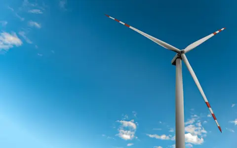 Mała elektrownia wiatrowa z białymi łopatami i czerwono-białymi końcówkami na tle błękitnego nieba z chmurami.