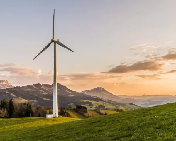 Wiatrak na zielonym wzgórzu, symbolizujący czystą energię z wiatru, na tle malowniczych gór o zachodzie słońca.