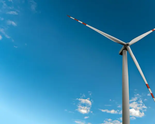Mała elektrownia wiatrowa z białymi łopatami i czerwono-białymi końcówkami na tle błękitnego nieba z chmurami.