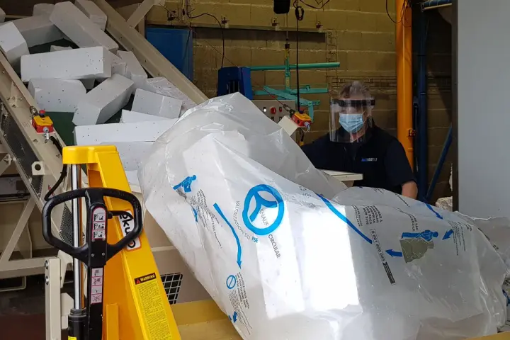 Pracownik w masce i przyłbicy przygotowuje styropian do recyklingu. Duży worek z logo Knauf Circular symbolizuje proces recyklingu styropianu.