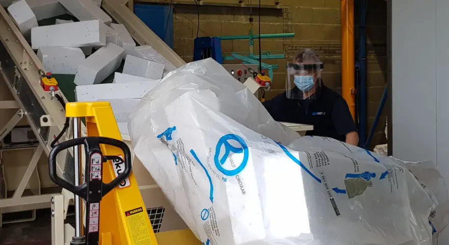 Pracownik w masce i przyłbicy przygotowuje styropian do recyklingu. Duży worek z logo Knauf Circular symbolizuje proces recyklingu styropianu.