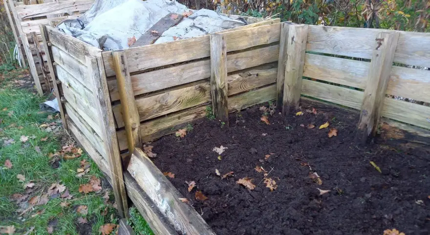 Drewniany kompostownik w ziemi, częściowo przykryty plandeką. Widać jesienne liście i trawę.