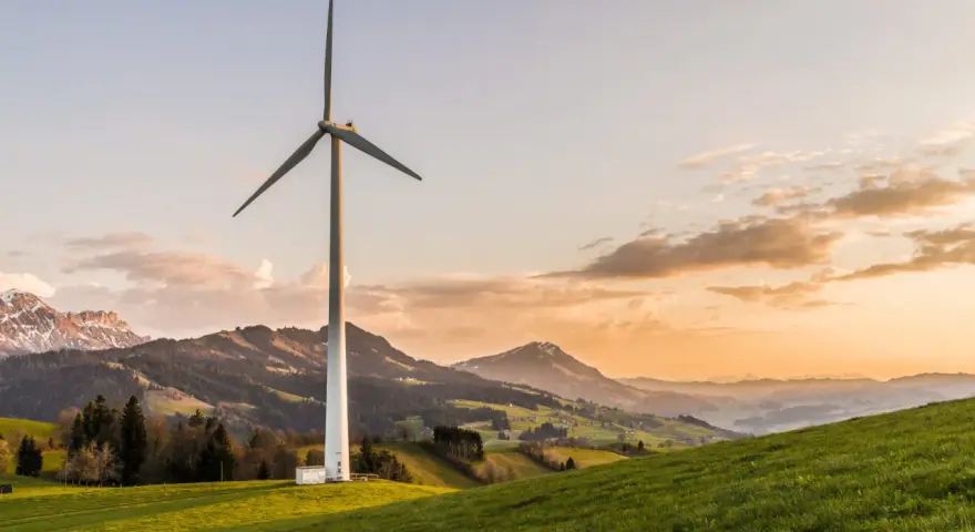 Wiatrak na zielonym wzgórzu, symbolizujący czystą energię z wiatru, na tle malowniczych gór o zachodzie słońca.