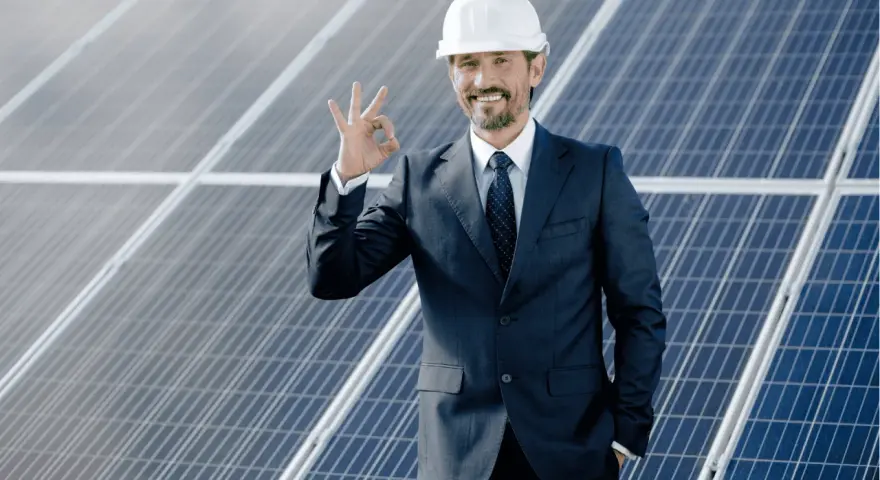 Uśmiechnięty mężczyzna w kasku i garniturze pokazuje znak OK na tle paneli fotowoltaicznych. To symbol efektywnego oddawania energii do sieci.