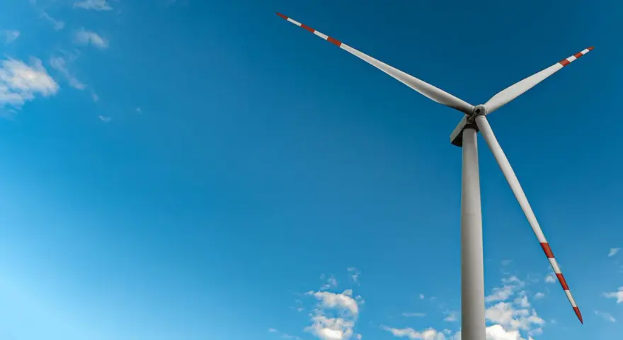 Mała elektrownia wiatrowa z białymi łopatami i czerwono-białymi końcówkami na tle błękitnego nieba z chmurami.