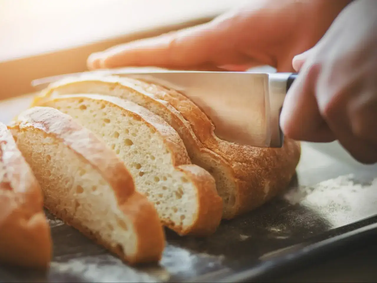 Ręka kroi nóżem czerstwy chleb na desce. Kromki są już pokrojone, a światło pada na chrupiącą skórkę.
