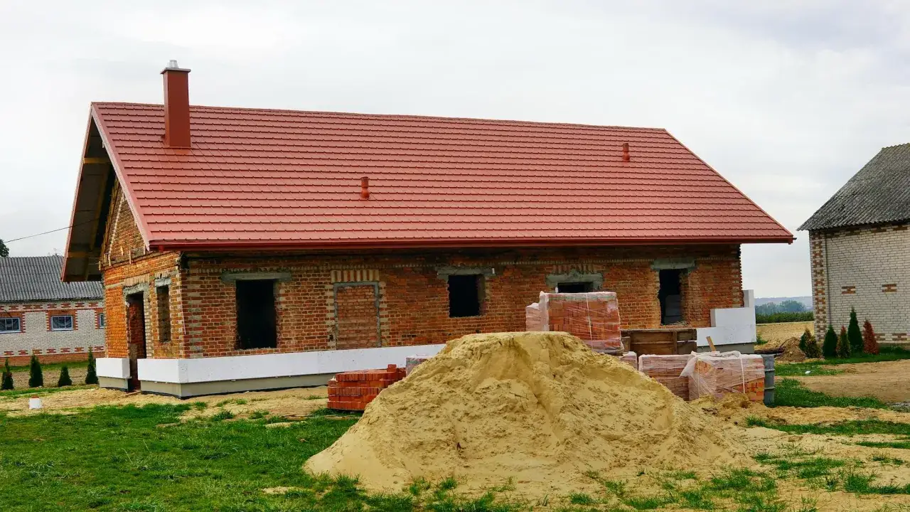 Budowa domu z czerwonej cegły, czerwony dach, komin, piasek i materiały budowlane. Wkrótce ulga na remont.