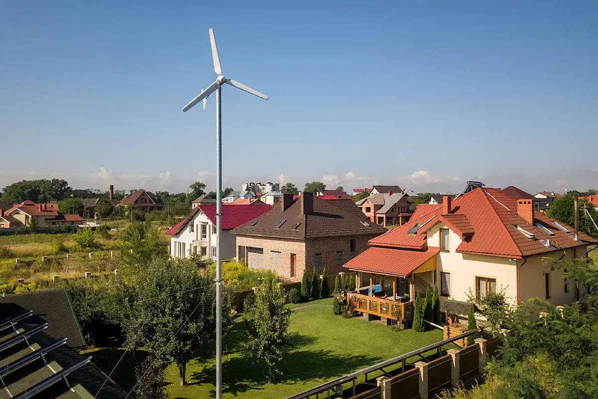 Wiatrak na podw&oacute;rku, być może turbina wiatrowa bez pozwolenia, na tle dom&oacute;w z czerwonymi dachami.