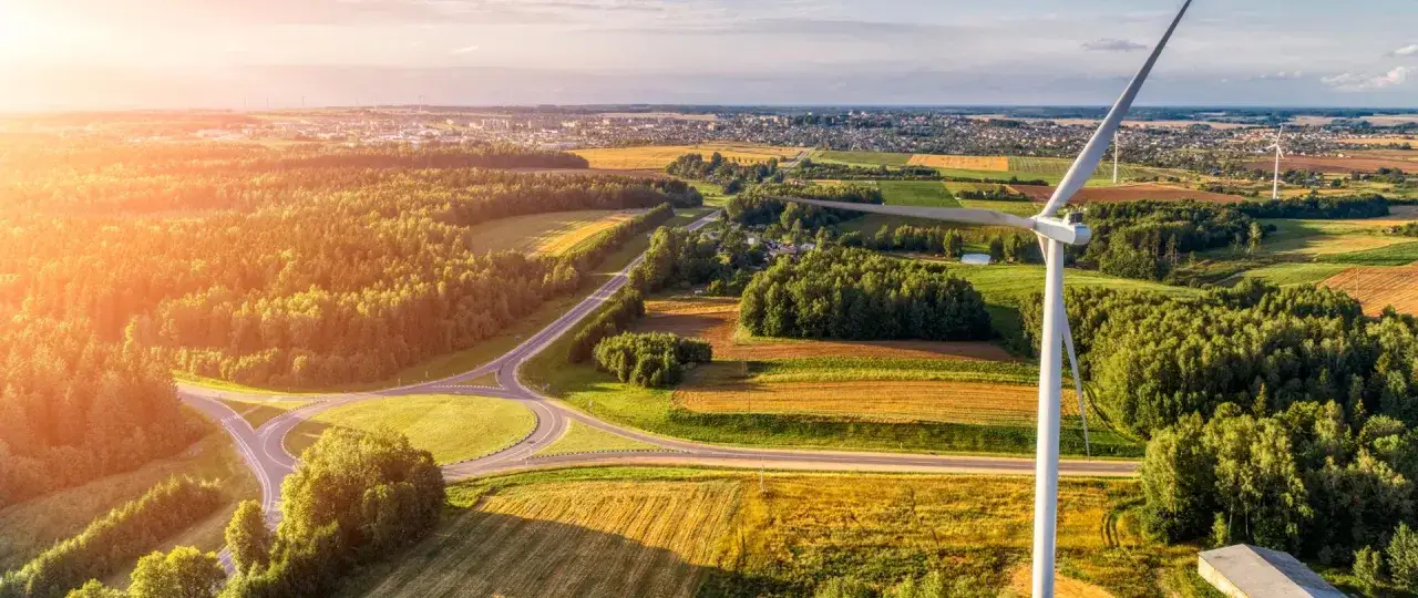 Wiatrak na tle pól i lasów, symbolizujący czystą energię z wiatru. Słońce zachodzi, malując niebo na pomarańczowo.