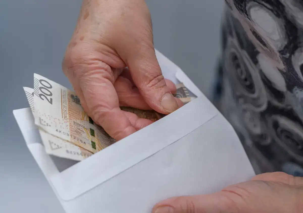 Dłoń wkłada banknoty 200 zł do koperty. Może to być związane ze złożeniem wniosku o zniżki na prąd dla emerytów.