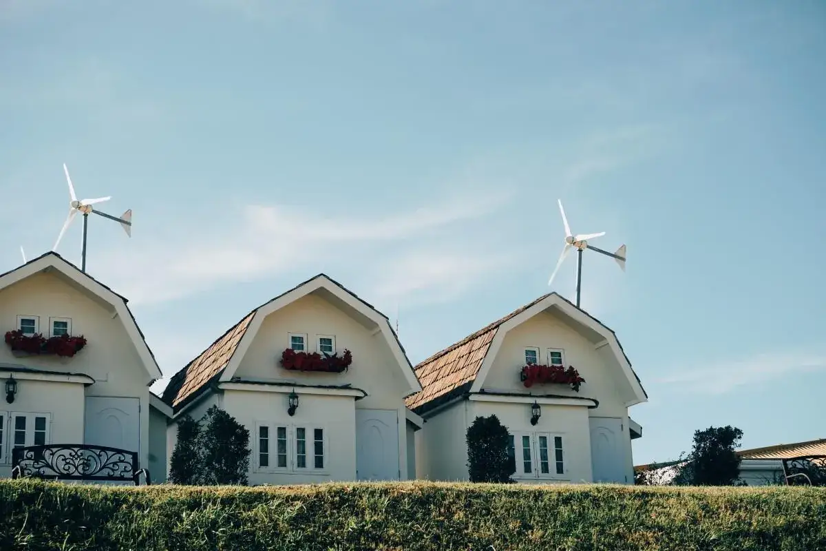 Trzy białe domki z czerwonymi kwiatami i turbina wiatrowa bez pozwolenia na dachu.