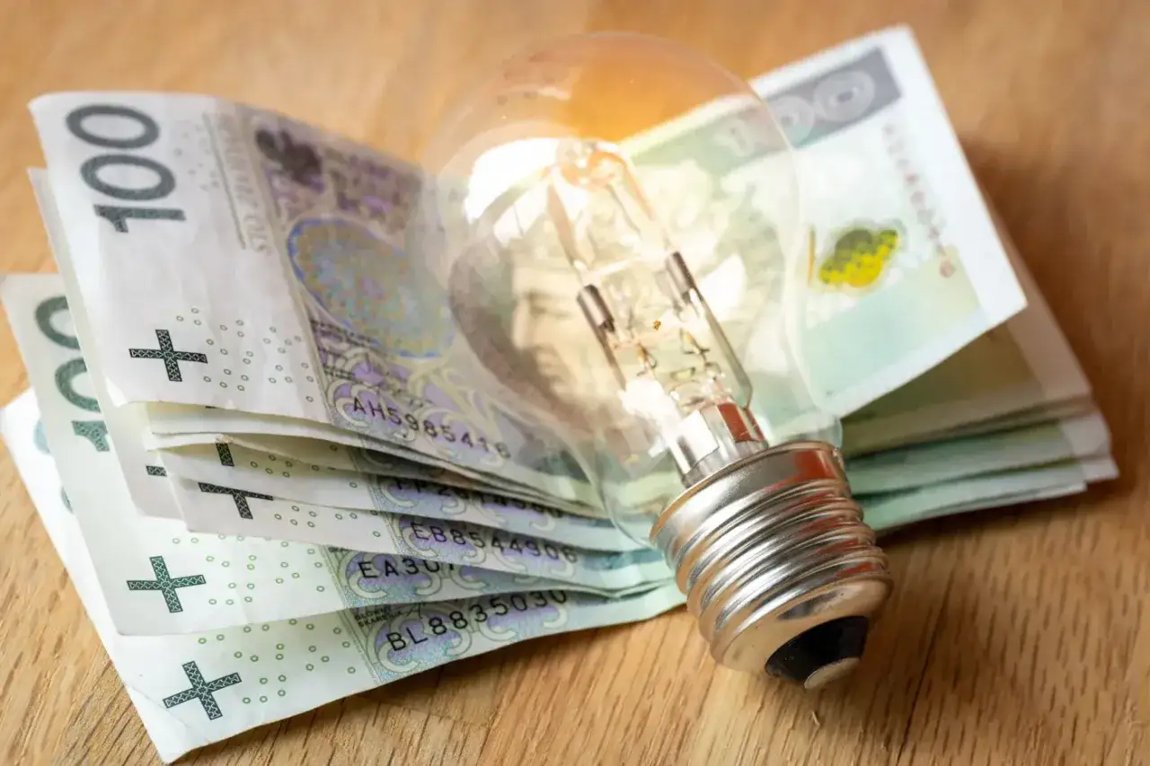 Żarówka na stosie banknotów 100 zł symbolizuje wysokie koszty energii i potrzebę dopłat do prądu.