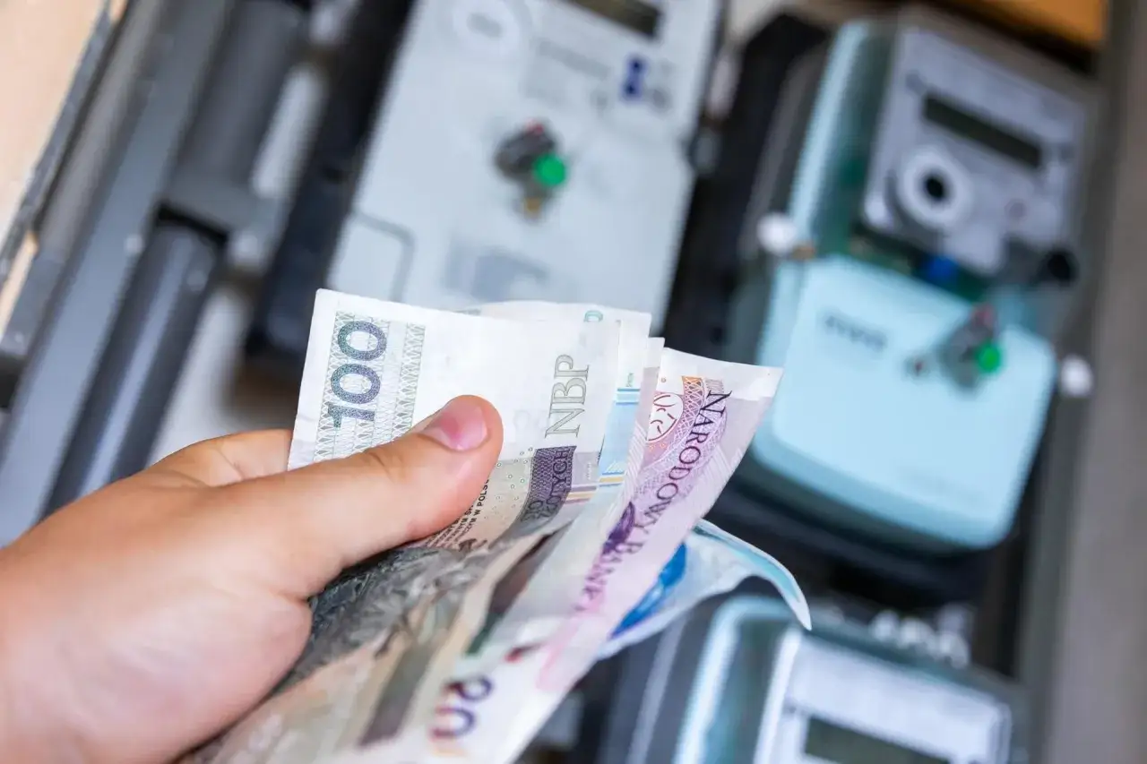 Dłoń trzyma polskie banknoty na tle liczników energii. Zastanawiasz się nad zniżki na prąd dla emerytów wniosek?