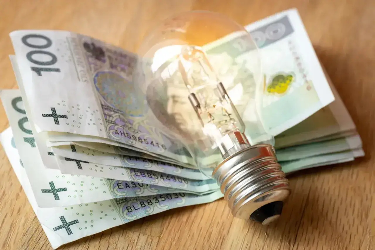 Żarówka na stosie banknotów 100 zł. Symbolizuje wysokie koszty energii, czyli ryczałt energetyczny.